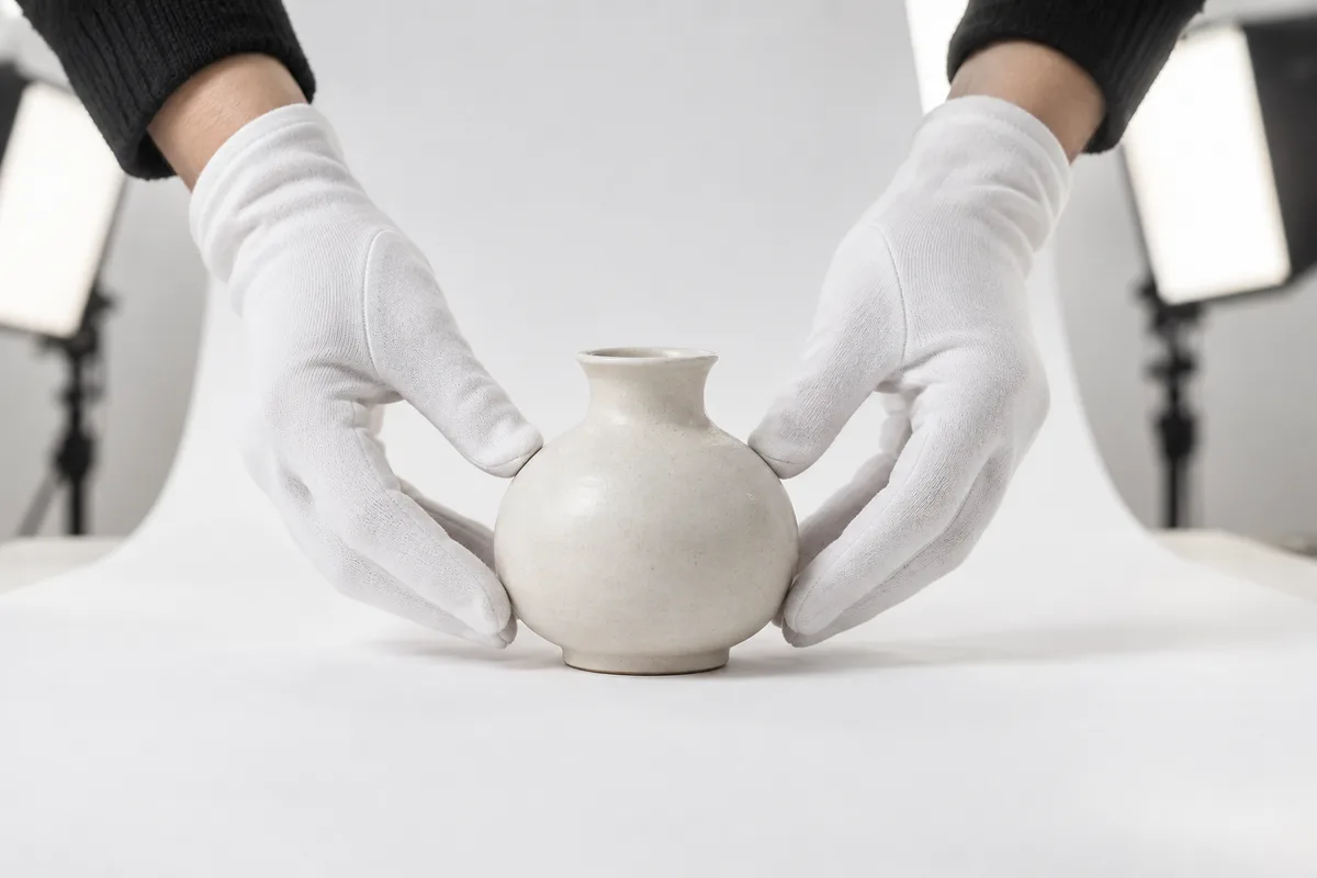Mains gantées disposant un objet en céramique claire sur un fond blanc de studio éclairé par deux boîtes à lumière